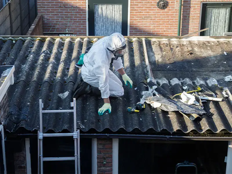 Worker in protective suit removing damaged roofing on a house.