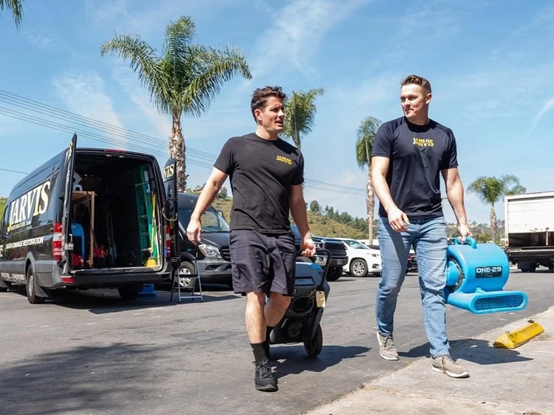 Two men carry equipment beside a service van in a parking lot.